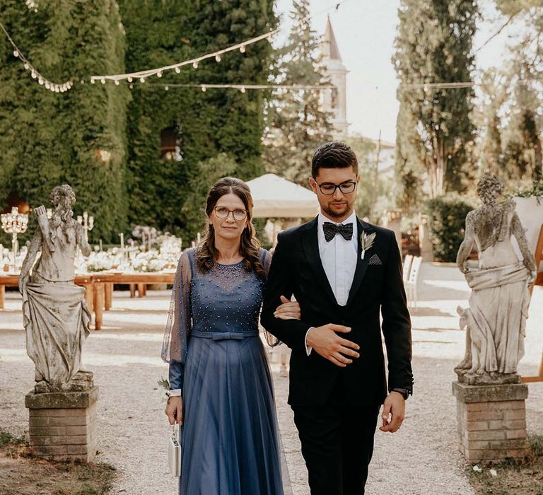 Groom walks down in the aisle with his mother who wears long deep blue gown | Royal Studio