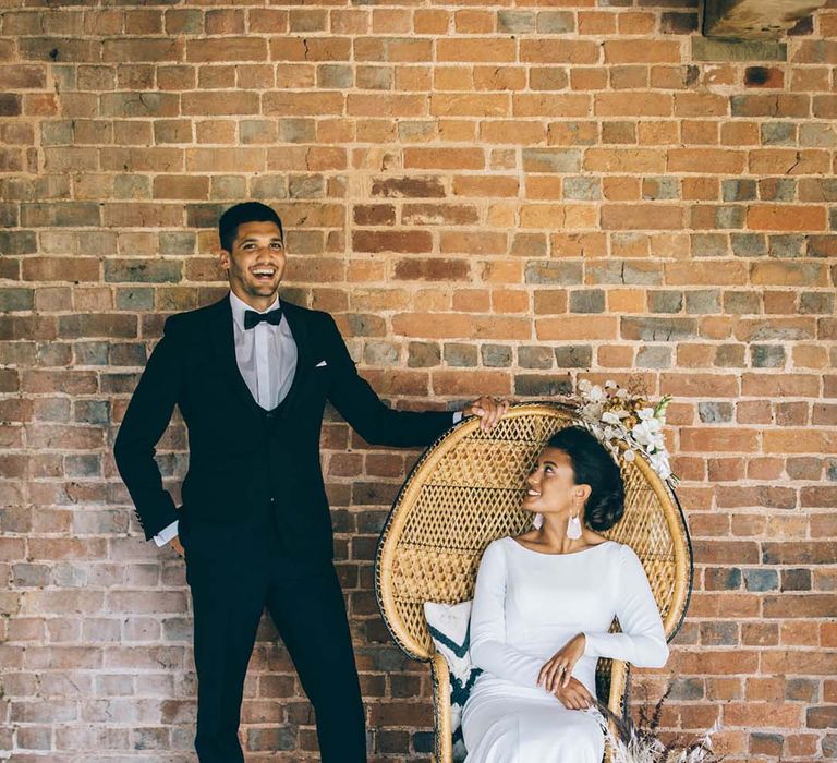 Boho black tie wedding with groom in a tuxedo and bride in a fitted wedding dress sitting on a wicker peacock chair 