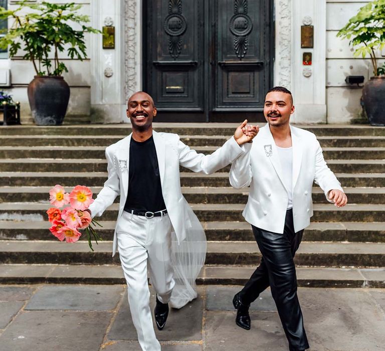 two stylish grooms in a white satin suit, cape, leather trousers and double breasted jacket holding hands at their city elopement 