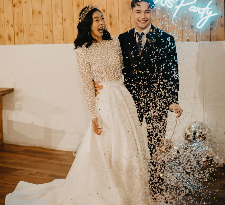 Bride in long sleeved applique wedding dress stands with groom in dark suit and floral tie as disco ball piñata confetti falls at rustic wedding with neon sign