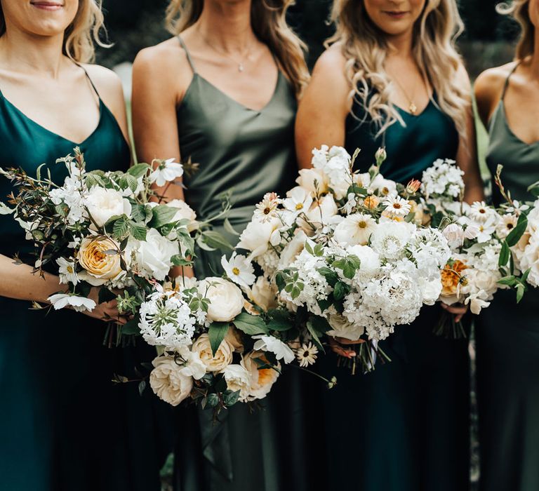 Bridesmaids in mixed green satin bridesmaid dresses hold white and green wedding bouquets at church wedding