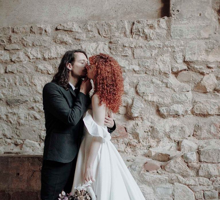 Groom in a wool suit kissing his bride in a princess wedding dress with red curly hair 