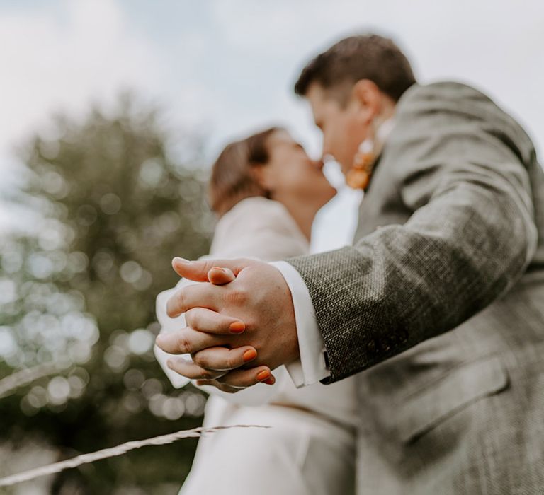 Groom in a grey check suit holding hands with his bride with orange nail polish 