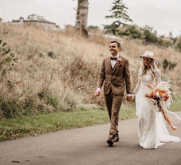 Groom in three piece brown tweed suit and bow tie walks down road holding hands with bride in two piece lace boho wedding outfit with bridal hat and white and orange boho bridal bouquet