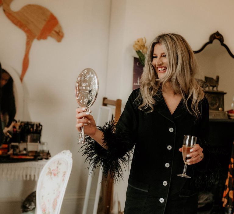 Smiling bride in black fur trimmed satin pyjamas holding glass of sparkling wine looks in handheld mirror before wedding