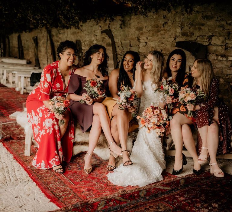 Bride in boho wedding dress sits on wooden bench with bridesmaids in mismatched bridesmaids dresses in stone barn