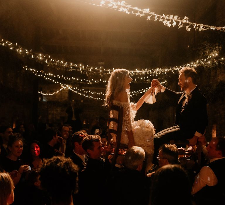 Laughing bride holds hands with groom during horah at evening wedding ceremony under fairy light canopy