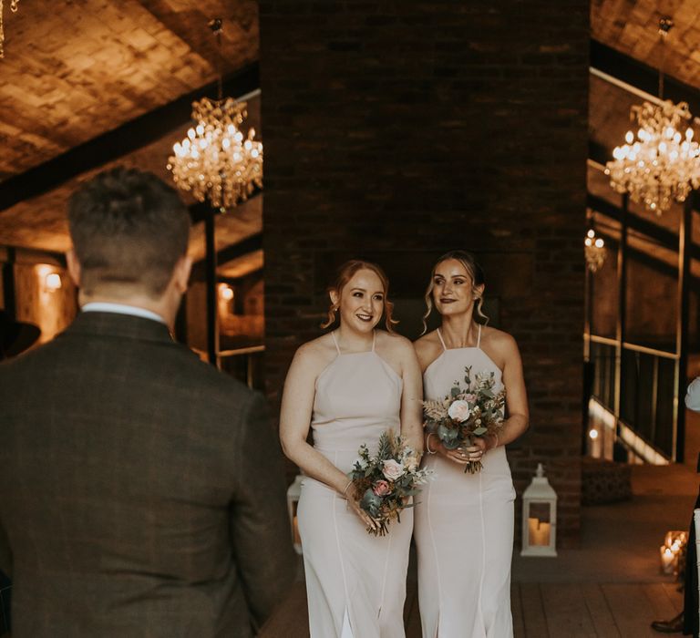 Two bridesmaids in light pink halterneck bridesmaid dresses holding mixed rustic bridal bouquets walk down barn aisle 