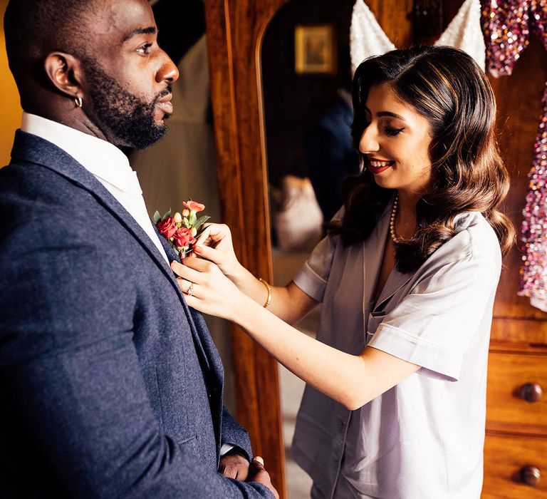 Bride in short sleeve pyjamas and pearl necklace fastens buttonhole of her bridesmaid wearing blue suit before her wedding