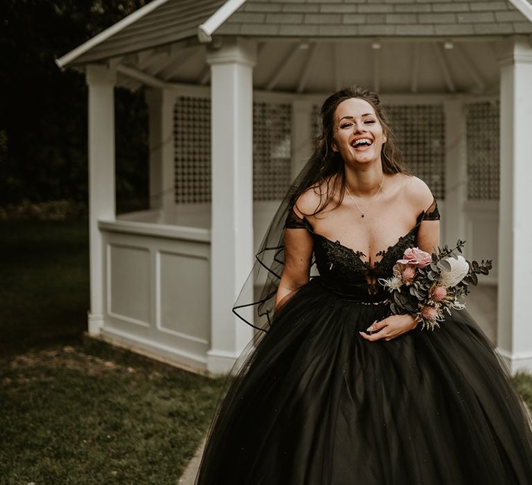 Bride laughs and wears black wedding gown with off the shoulder straps and holds floral bouquet 