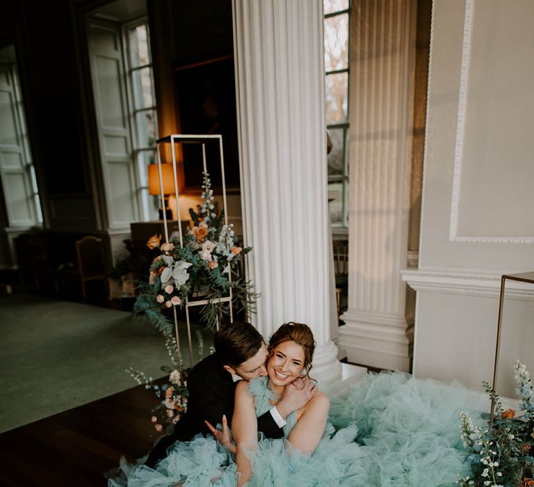 bride in a teal wedding dress sitting on the floor being kissed by her husband in a tuxedo surrounded by luxury floral arrangements 