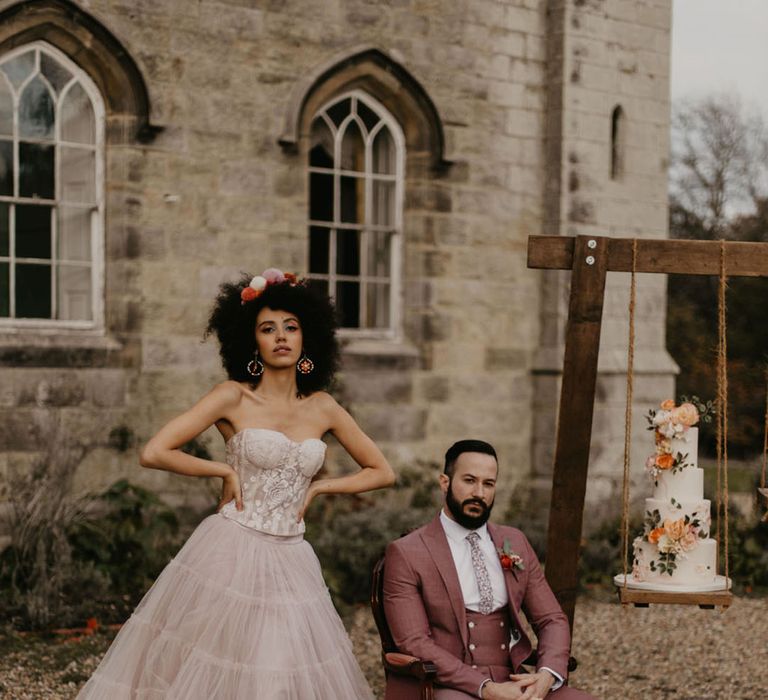 Black bride with afro hair in a pink tulle skirt and corset top standing next to her groom in a pink three-piece suit and wedding cake on a swing 