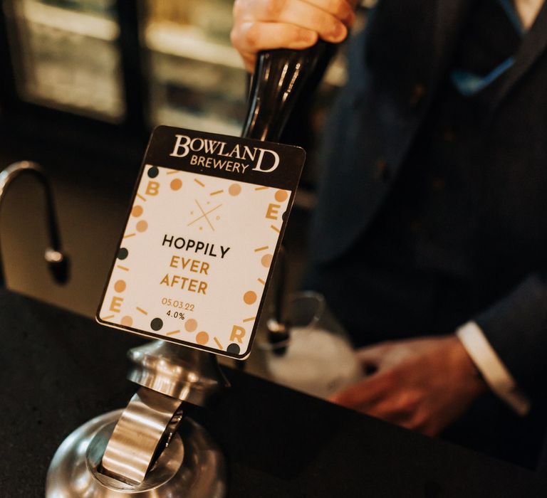 Groom in navy suit pulls a draft ale from the pump labelled 'Hoppily Ever After' during wedding reception at Holmes Mill