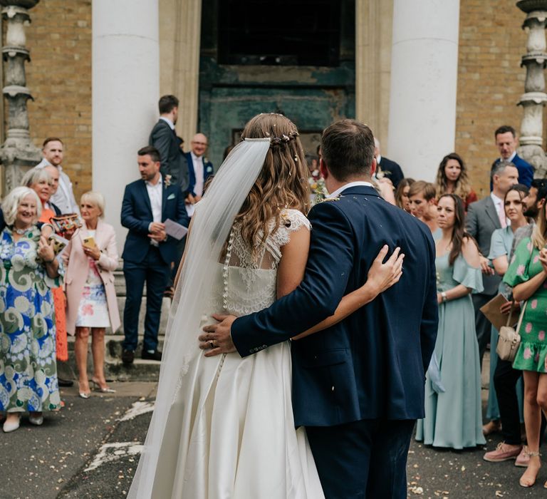 Bride with waterfall braid hairstyle and buttoned lace wedding top and pleated skirt standing with groom in navy blue 