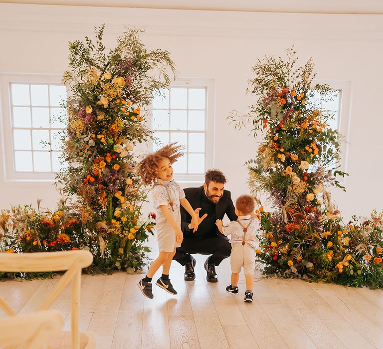 Groom in all-black playing with page boys in floral shirts, braces and shorts in-between orange and green column wedding ceremony flower arrangements 