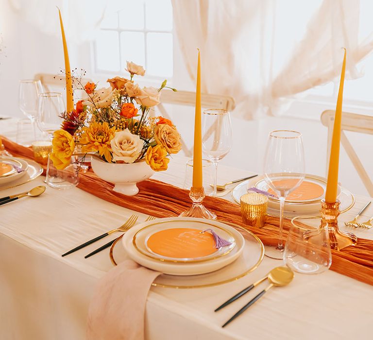 golden wedding table decor with orange table runner, yellow taper candles and flower arrangements 