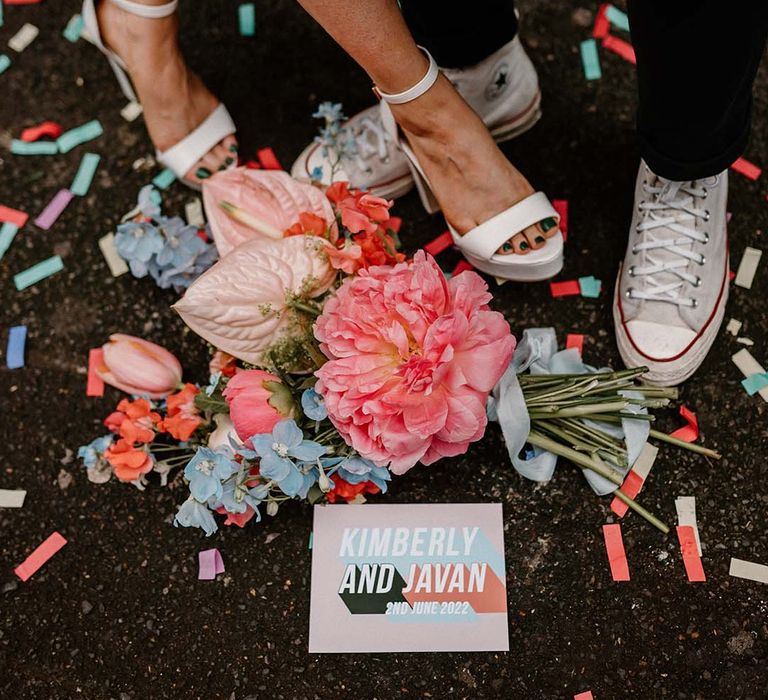 Coral pink peony, baby pink anthuriums and blue delphinium wedding bouquet resting on a floor sprinkled with confetti 