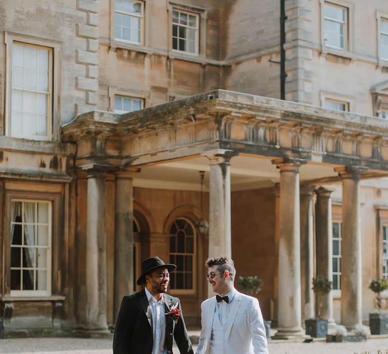 Groom in a two piece black suit, fedora hat and boots holding hands with his partner in a white three-piece groom suit and white boots at Prestwold Hall Barns