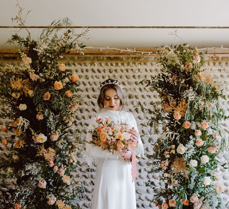 Bride in a stain wedding dress with long sleeves and tassels standing at the altar of Montague Farm Hankham between two column floral arrangements 