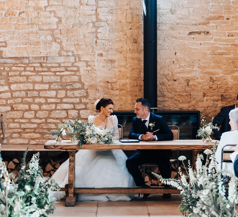 Bride and groom sit at wooden table under a disco ball as they sign their wedding register 