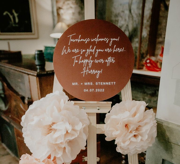 Small brown circular welcome wedding sign on wooden easel decorated with pink and peach flowers