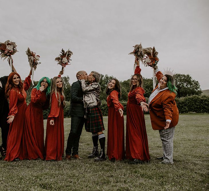 Grooms kiss in the middle of their bridesmaids who wear deep red gowns from ASOS and hold homemade floral bouquets 