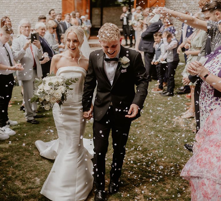 Groom in black tie and bride in strapless white wedding dress have their confetti exit to white petals 