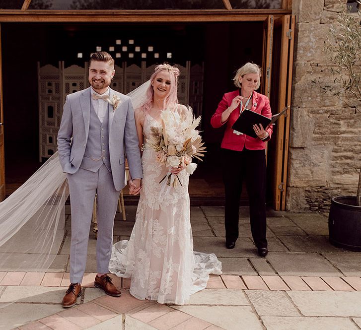 Bride wearing floral bridal crown and holding large dried floral bouquet featuring pampas grass stands with her groom in pale grey three piece suit