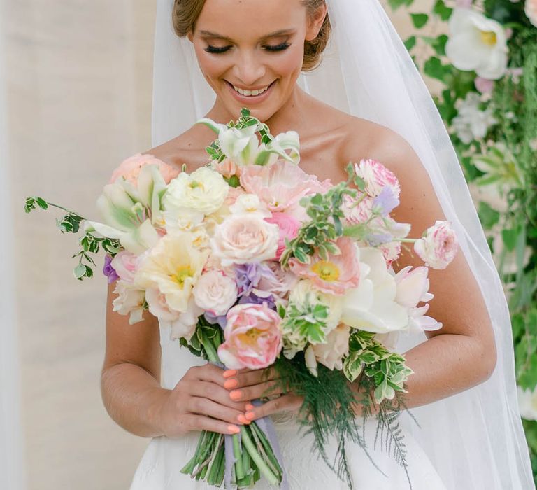 Bride wearing a beaded pearl headband smiles as she looks at her pretty bridal bouquet of pink, white and purple flowers with roses and lilies 