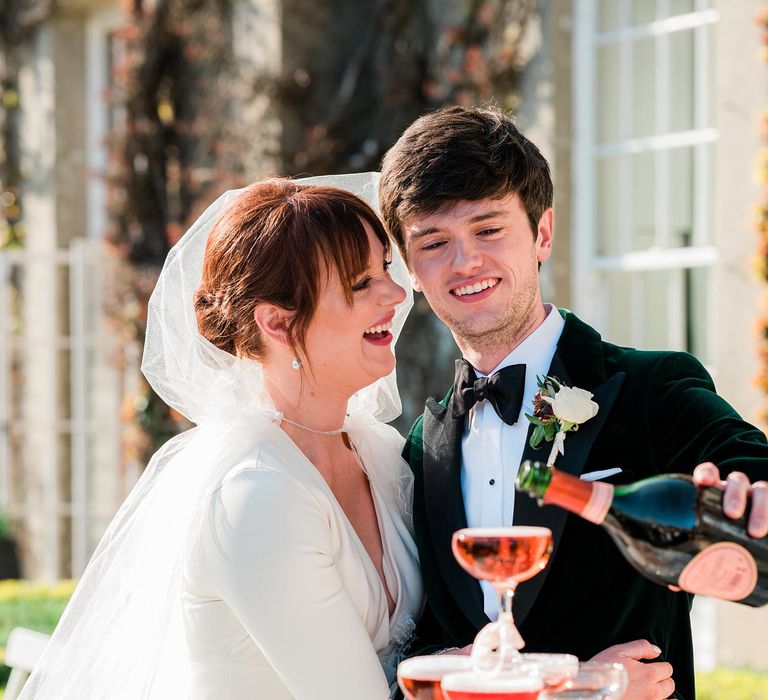 Groom pours champagne into champagne tower as bride wears hood styled cathedral veil on their wedding day at Findon Place 