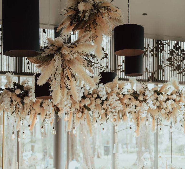 Pampas grass installations hang above banquet tables complete with festoon lighting and pastel pink fabric table runner 