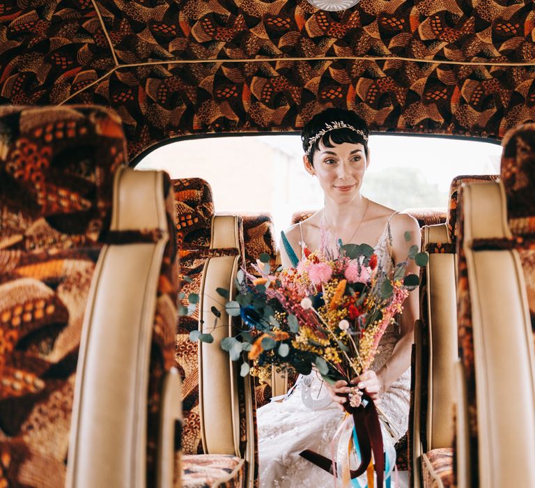 Bride holding brightly coloured floral bouquet sits in vintage bus and wears embellished bridal crown