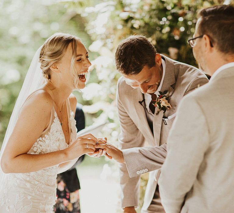 Bride laughs wearing a lace wedding dress and veil as he she puts on the groom's wedding band during the ceremony 