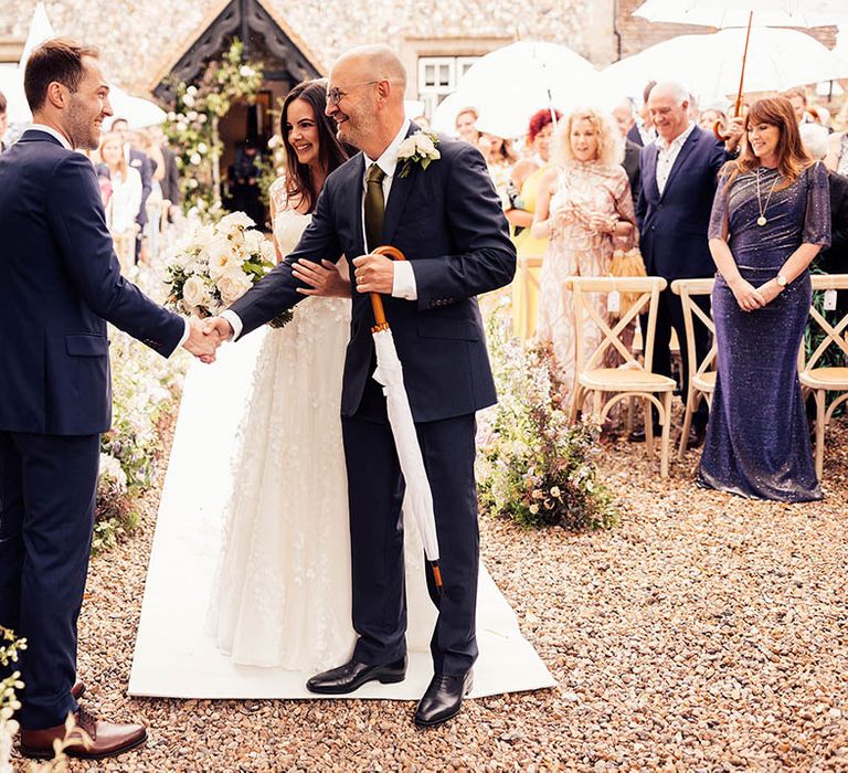 Father of the bride shakes the groom's hand at the end of the aisle with the bride on his arm 