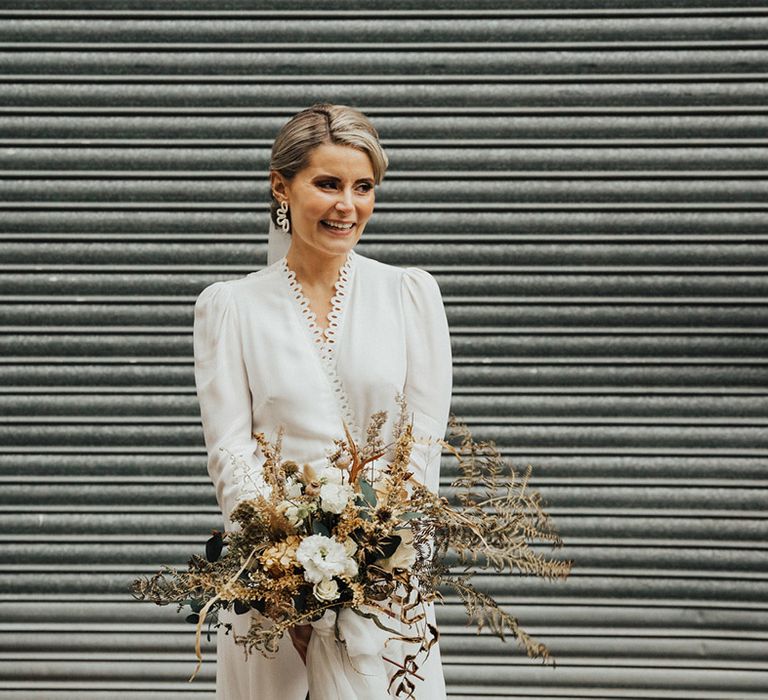 Bride wears Reformation wedding dress with scalloped lace edge to neckline whilst holding dried floral bouquet 