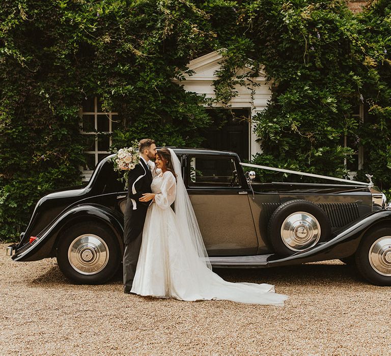 Bride and groom stand next to their classic black vintage wedding car outside of their country house wedding venue 