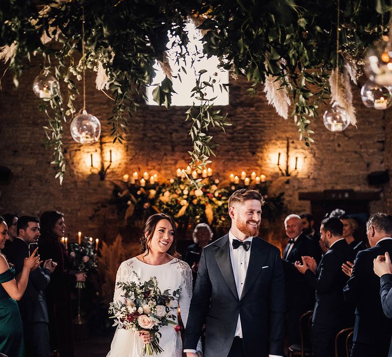 Smiling groom in a custom black tie suit and the bride in Atelier Pronovias exit their ceremony as a married couple 