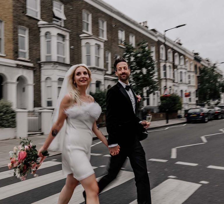 Bride in white short satin dress with feather trimmed neckline and groom in black tux with bowtie walking across the road 