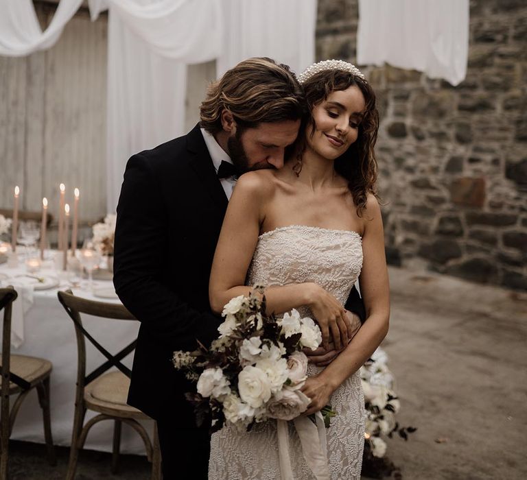 Bride in sleeveless lace wedding dress wearing a pearl headband and silver belt standing with groom in black tux