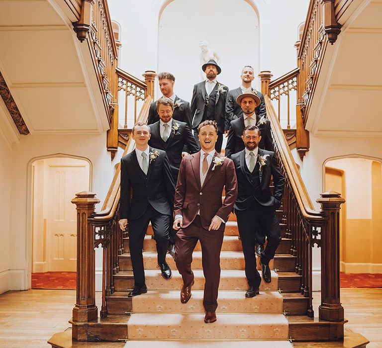 Groom in maroon suit walks down staircase with his groomsmen in blue suits 