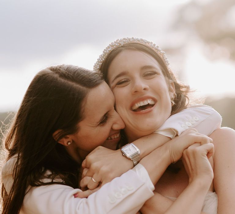 The bride in a white suit jacket and silver watch hugs the bride in a pearl headband and off the shoulder wedding dress from behind 