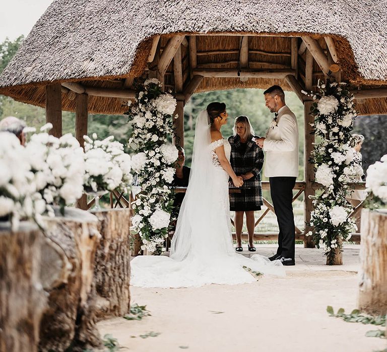 The groom reads out his vows to the bride as they stand next to white flower columns for their outdoor wedding ceremony  