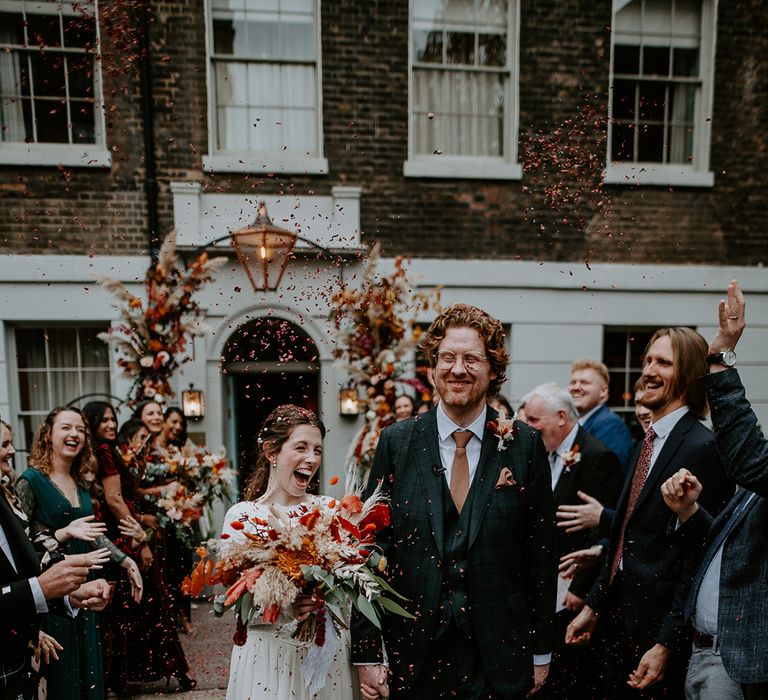 Bride in long sleeve lace vintage wedding dress and closed toe satin heels holding large autumnal bouquet with orange, black, green and yellow roses, peonies, foliage and pampas grass with groom in dark suit with orange tie and autumnal boutonniere doing confetti walk outside The Zetter Townhouse