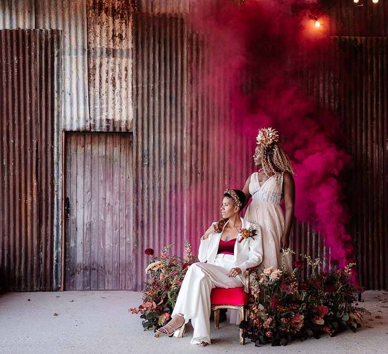 Bride in sleeveless floaty peach gown with embroidered stairs and gold leaf crown and bride in elegant white silk suit with autumnal dried floral boutonniere sitting on gold and red velvet chair with deep pink smoke bomb and dried autumnal arrangement