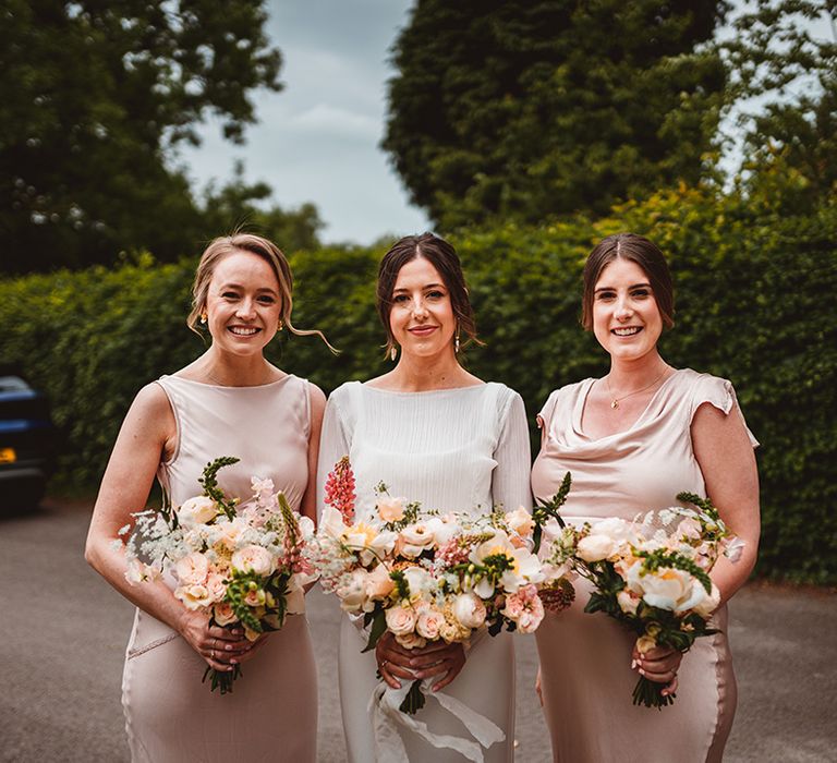 Bride in sheer bridal cover up top with bridesmaids in light pink satin bridesmaid dresses, one with cowl neck, the other high neck, all holding tropical wedding bouquets