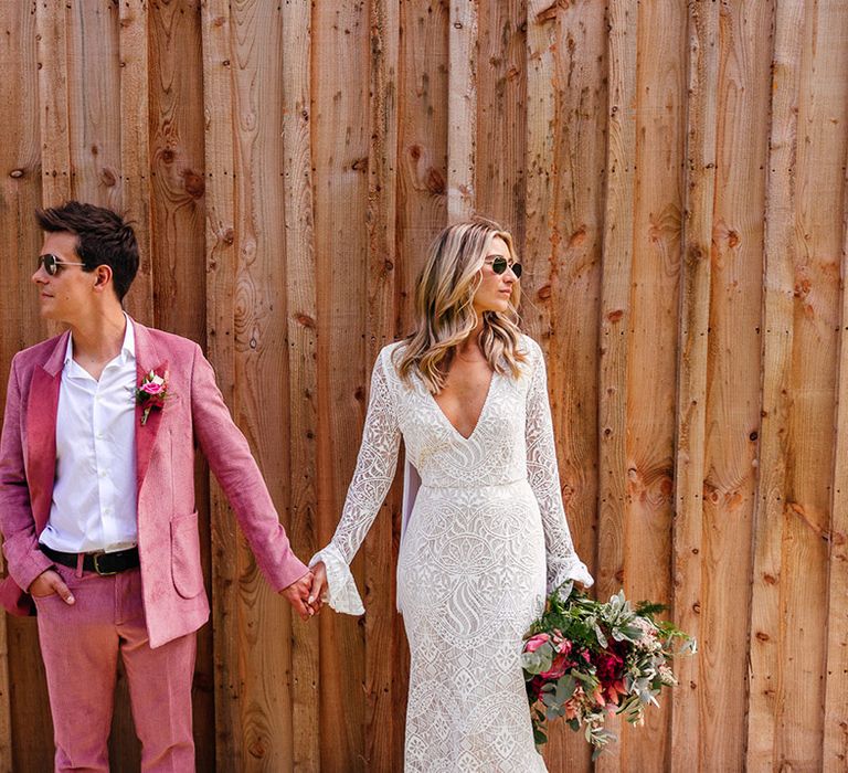 Bride in v neck long sleeve lace boho wedding dress with tassels and sunglasses holding hands with groom in pink corduroy suit with pink and red rose, foliage and dried flower boutonniere and sunglasses 