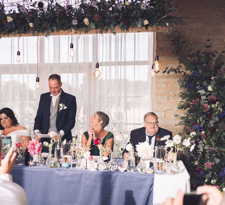 Groom in navy morning suit standing up giving a speech with the wedding party and guests sitting down at the rustic Stratton Court Barn venue 