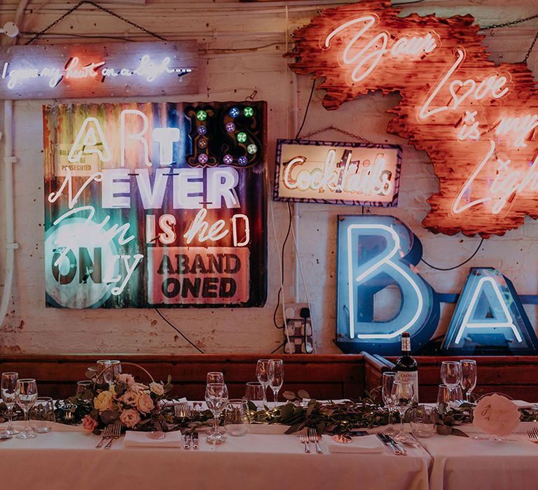 Rustic wedding tablescape with foliage and white rose table runners with colourful neon signs behind 