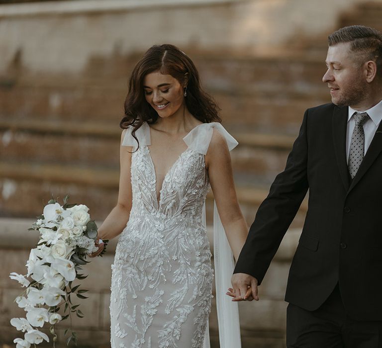 The bride in a fitted wedding dress with bow tulle straps with white cascading bouquet with groom in black suit 