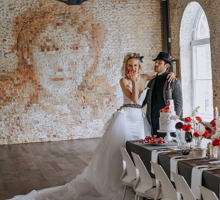 Bride in  romantic organza and tulle a-line wedding day bridal ballgown with a flowing skirt, sculpted bandeau top and patterned black belt eating a piece of cake with groom in dark grey jacket, black ribbed turtle neck, black trousers and a black fedora hat 